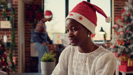 Canvas Print - African american festive woman browsing data on laptop, wearing santa hat at work in office during christmas holiday season. Employee celebrating winter tradition with decorations and working.