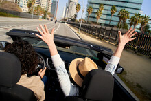 Cheerful Caucasian Couple Driving On Sunny Day In Convertible Car Enjoying The Freedom In A City. Touristic City Road Trip On A Country. Urban Landscape Holidays Happy Man And Woman Having Fun