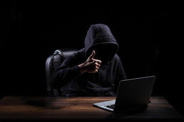 A hacker, dressed in a black hood, is raising a thumbs up when completing tasks to hack into a darkened room.