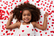Smiling Young Girl With Afro Hair Lying On Bed Surrounded By Heart Shaped Confetti