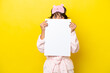 Young latin woman in pajamas isolated on yellow background holding an empty placard and hiding behind it