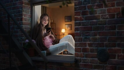 Poster - A Beautiful White Woman is Using her Smartphone While Sitting on her Windowsill at Night With a Fleece Blanket. Young Female Smiling While Browsing Through Potential Matches in an Online Dating App