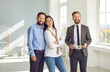© Studio Romantic - Young family decides to buy a house after a house tour. Portrait of a happy young couple together with a real estate agent standing in a big light room, looking at the camera and smiling