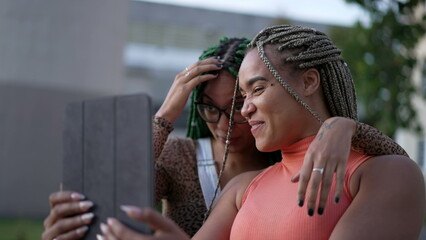 Two happy black adult girls talking to friend on tablet video. Young women with box braids hairstyle holding device outdoors in urban street. Real life authentic smile and laugh