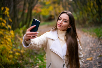 Wall Mural - Pretty young lady making selfie in autumn park. Beautiful attractive woman with phone.