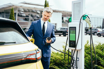 Handsome mature grey haired man holding charging cable at electric charging station point standing near his car outdoor.