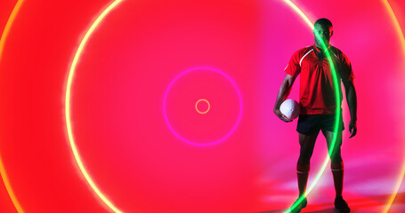 Sticker - African american rugby player holding ball over colorful illuminated circles on pink background