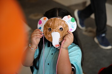 A little child girl wearing a colored animal paper mask fronting her face is isolated in the classroom background with an orange balloon. elephant paper mask art and crafts.