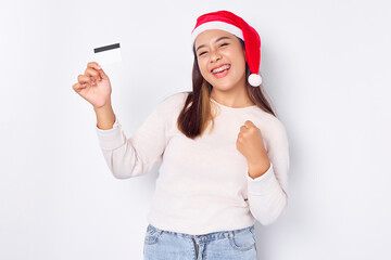 Excited young Asian woman in Christmas hat holding credit card and making success gesture isolated on white background