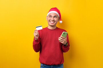 Smiling young Asian man in Santa hat using mobile phone and credit card, looking up, over yellow studio background. celebration Christmas holiday and New Year concept