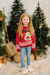 © Serhii - Child girl in stylish Christmas sweater with deer and comfortable home slippers near Xmas Tree. Smiling kid stands near Christmassy gift, presents for New Year. Decorated interior with garlands, light