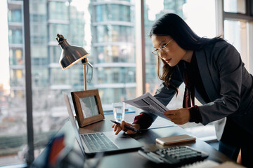 Wall Mural - Asian female CEO working on laptop while analyzing business reports in office.