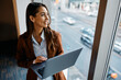 © Drazen - Happy female entrepreneur using laptop and looking through window in office.