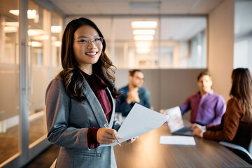 Happy Asian executive manager analyzing business reports with her team in office and looking at camera.