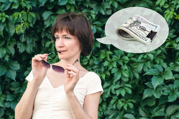 Woman with dark red hair stands and holds sunglasses in her hands n front of a wall covered in leaves. Woman bit her glasses and looks away. Woman thinks about money, salary or winning the lottery.