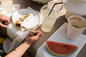 Young man eats cake with a golden spoon. only the hands visible. White plate with golden edges. Next to it is a drink with a straw, a cup of coffee and a piece of watermelon. Sunny day, outdoor party