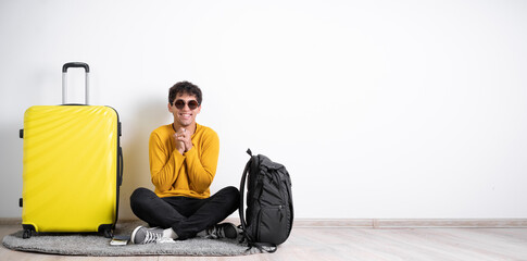 Wall Mural - Young guy traveler with a suitcase satisfied with a positive expression sits isolated on a white background holds his hands near his chest