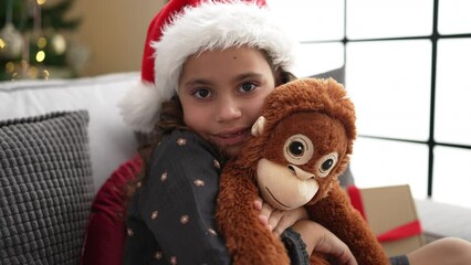 Sticker - Adorable hispanic girl hugging monkey doll sitting on sofa by christmas tree at home