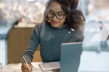 Wall Mural - Smiling woman freelancer working laptop and making notes while sitting near windows in cozy cafe