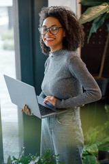 African businesswoman holding laptop and looking at camera while standing near window in coworking