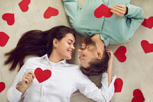 Happy Beautiful Couple Spending St Valentine's Day Together. Young Man And Woman Lying On Floor Among Red Heart Shaped Cards, Looking At Each Other And Smiling. Top View, High Angle Shot, From Above