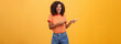 Ask him. Portrait of friendly and joyful good-looking stylish female shop assistant with curly hair and dark skin pointing left with both hands, smiling assured and entertained over orange background