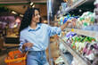 © Liubomir - A young beautiful Hispanic woman buys cherry tomatoes in a supermarket. She stands smiling near the refrigerator, the vegetable department with a basket for groceries.