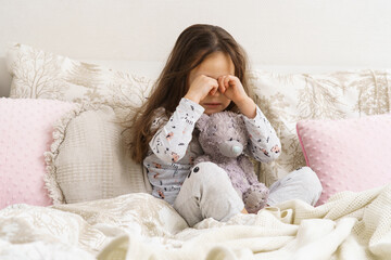 Depressed, sad, disappointed kid, girl rubbing eyes, sit with stuffed bear toy in soft white bed in the morning. Abandoned, orphan children, sadness and stress. Painful emotions and loneliness