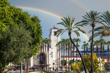 Union Station train station surrounded by office buildings, parked cars, tall lush green palm trees and plants and people walking on the sidewalk with blue sky and a rainbow in Los Angeles California