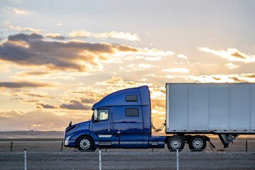 Profile of big rig blue semi truck tractor with dry van semi trailer driving on the flat highway road along the fields at twilight