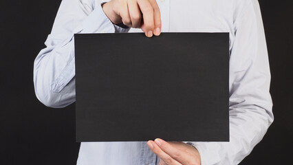 Wall Mural - Empty black board A4 paper in man hand and wear blue shirt on black background.Crop body part.