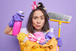 © wayhome.studio  - Serious Asian housewife holds cleaning detergent and broom stares impressed wears rubber gloves collects laundry in basket isolated over purple background. Domestic chores and housework concept