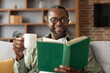 Happy mature black guy in glasses enjoys cup of coffee and reads book in free time in living room interior, close up