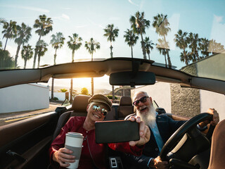 Senior couple having fun doing selfie inside a convertible car during holiday vacation - Elderly travel concept