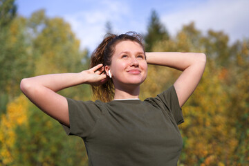 Wall Mural - portrait of beautiful happy girl, young positive woman standing in golden autumn forest or park, listening to music in wireless earphones smiling on natural background, breathing deep deeply fresh air