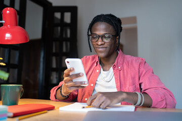 Happy smiling African student guy looking at smartphone screen chatting with girlfriend while studying at home, joyful black man sitting at desk using phone during study. Mobile distraction concept