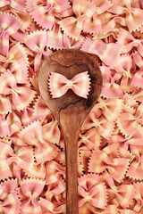 Wall Mural - Farfalle bow tie Italian pasta background with olive wood spoon. Beetroot food dye used for pink colour. Healthy gourmet eating concept. Flat lay top view.