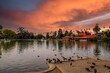 a gorgeous autumn landscape at Lincoln Park with a lake surrounded by lush green palm trees and plant, an orange building with red brick, birds on the banks, powerful clouds at sunset in Los Angeles