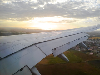 Airplane window view of the aircraft wing with a cloudy sky with golden hour sunlight at sunset. Algiers city great mosque minaret visible from far away. Houses and buildings with agricultural fields.