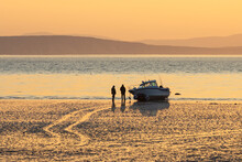 Boat On The Shallows At Low Tide. Two Men Near The Boat Are Waiting For The Tide. Morning Seascape At Sunrise. View Of The Shallows And The Sea Bay. Golden Morning Sunlight At Dawn. Natural Background