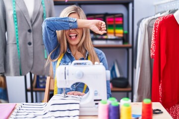 Poster - Blonde woman dressmaker designer using sew machine smiling cheerful playing peek a boo with hands showing face. surprised and exited