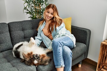 Sticker - Young caucasian woman sitting on sofa playing with dog at home