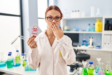 Wall Mural - Young caucasian woman working at scientist laboratory holding toxic label covering mouth with hand, shocked and afraid for mistake. surprised expression