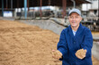 Positive man farmer standing at large heap of brewer's spent grain and holding bunch in hands.