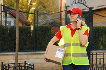 Canvas Print - Courier in uniform with parcel talking on smartphone near private house outdoors, space for text