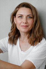 Closeup portrait of happy creative young woman smiling and looking directly to the camera over grey wall. Female at her 40s relaxed in white t-shirt