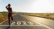 New Year 2023 Or Start Straight Concept.word 2023 Written On The Asphalt Road And Athlete Woman Runner Stretching Leg Preparing For New Year At Sunset.Concept Of Challenge Or Career Path And Change.