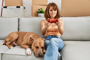 Poster - Young caucasian woman playing video game sitting on sofa with dog at home