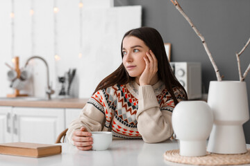 Sticker - Young woman with cup of hot tea at table in kitchen