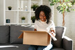 © fotofabrika - Young african woman sit on couch at home unpacking parcel cardboard box with online purchase